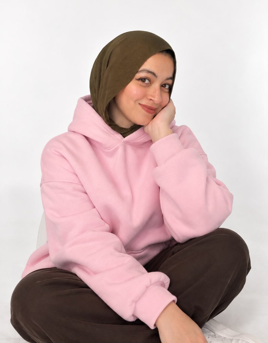 Salma Osama sitting cross-legged in a pink hoodie and olive hijab, smiling with her chin on her hand